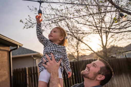 Young Father Holds Up Two-year Old daughter as she touches an edison light bulb decoratively arranged in a back yard at dusk
